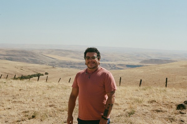 Person with short dark hair and a rust red shirt in an open landscape with yellow grass fields