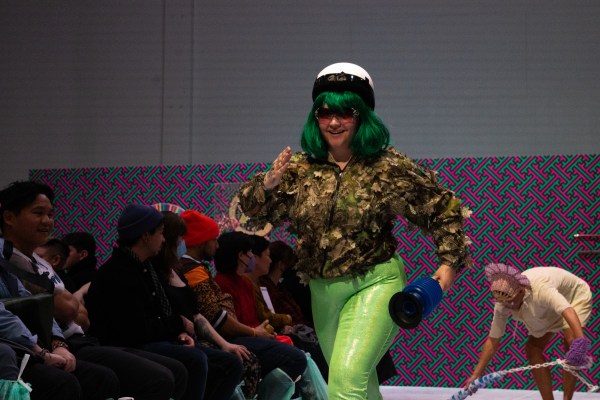 central figure with lime green pants, a camouflage jacket, white helmet and sunglasses. Figure appears to be in a running motion. Figures in the audience behind