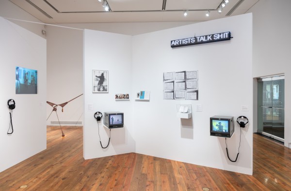 gallery install view with white walls, television screens with headphones and various ephemera. On the upper right corner of the gallery wall is an led sign that reads 'artists talk shit'